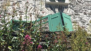 Un bâtiment en pierre doté d’une tour, d’une fenêtre avec des volets verts et entouré de végétation.
