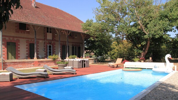 A swimming pool with lounge chairs, a covered patio, and a house with a red-tiled roof.
