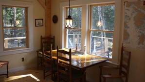 Un coin repas avec une table et des chaises en bois, de grandes fenêtres donnant sur la verdure, un luminaire suspendu et une carte accrochée au mur.