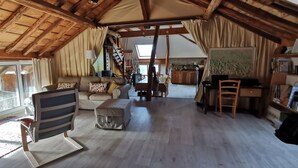 A spacious loft-style room with a high ceiling, wooden beams, a large window, a sofa, a dining table, and a kitchen area.
