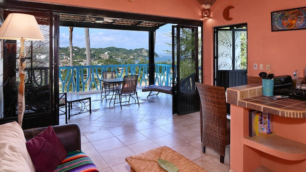 Un salón acogedor con sofá, una mesita y un banco de mimbre. Hay un balcón con vistas al mar y las colinas.