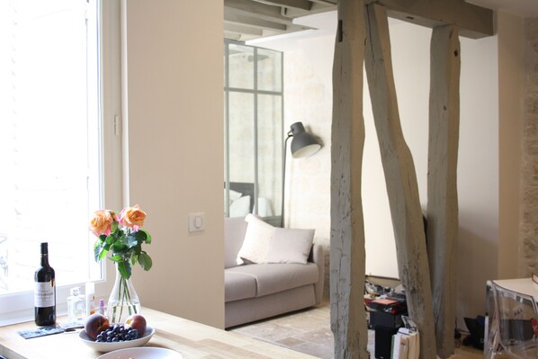 A room with a wooden beam ceiling, a window with a view, a sofa, and a table with a vase of flowers and a bowl of fruit.