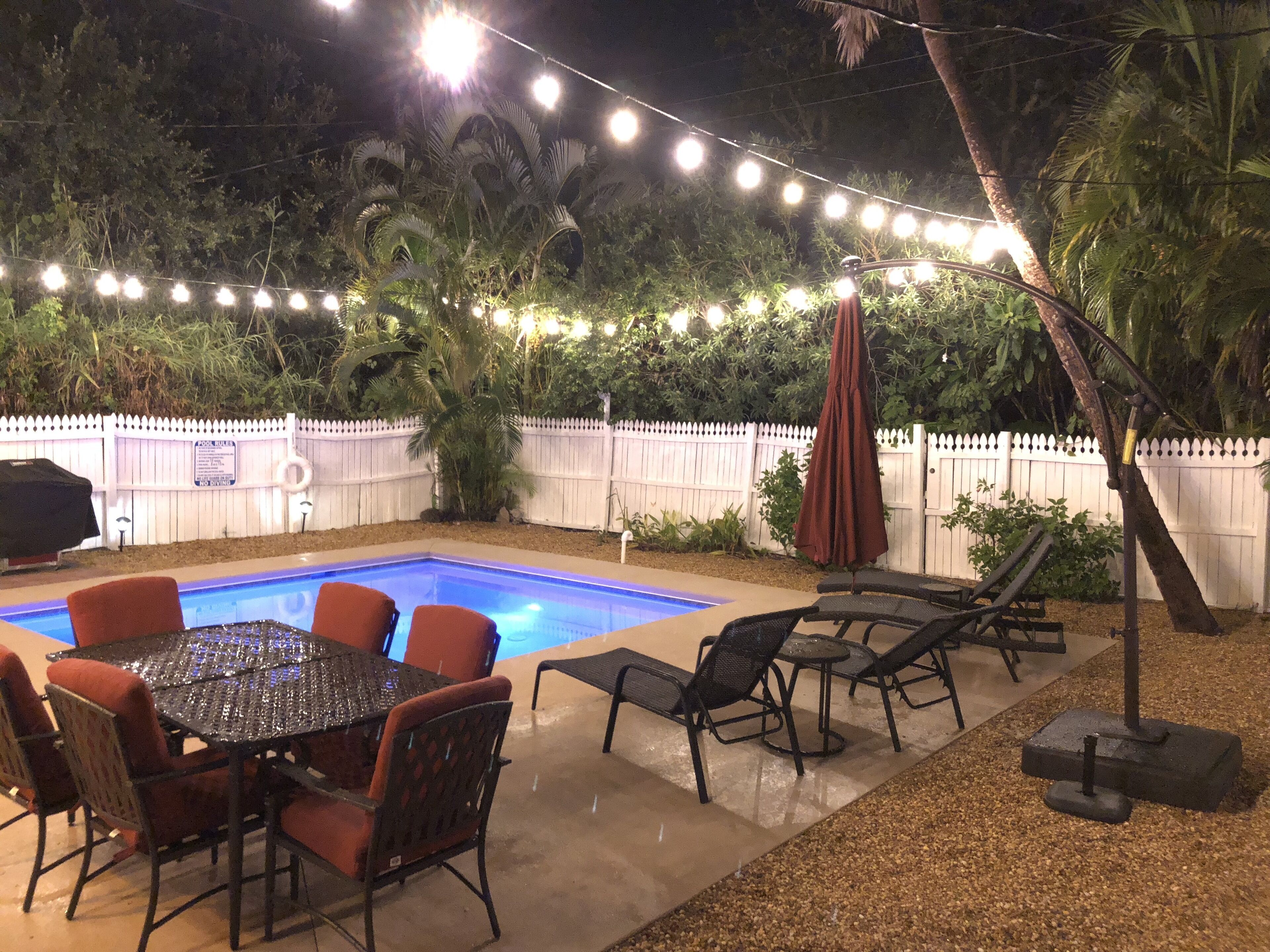 A backyard with a swimming pool, outdoor seating, string lights, and a white picket fence.