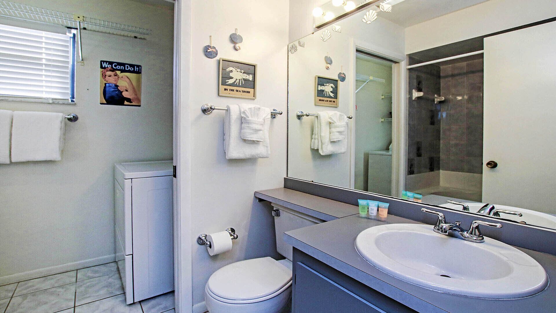 Une salle de bain moderne avec un grand miroir, des éléments d’ameublement blancs et une affiche accrochée au mur.