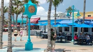 Zona turística tropical con palmeras, mobiliario exterior y un reloj turquesa.