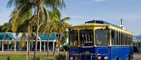 Un autobús azul y amarillo antiguo estacionado en una zona pavimentada, con palmeras y un edificio al fondo.