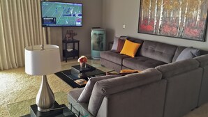 Ett modernt vardagsrum med en grå soffa, ett soffbord med en lampa, en tv på väggen och en tavla.