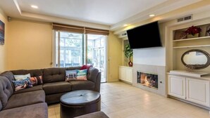 A living room with a sectional sofa, a round coffee table, a fireplace, a flat-screen TV mounted on the wall, and a large window with blinds.