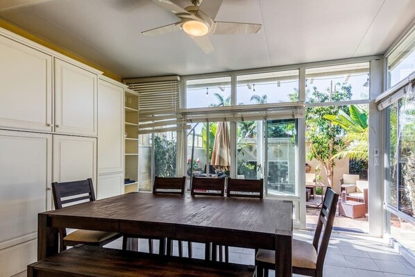 Uma área de refeições com mesa e cadeiras de madeira, janelas amplas e um ventilador de teto.