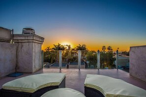 Terraço no telhado com duas cadeiras de descanso e vista do pôr do sol sobre palmeiras.