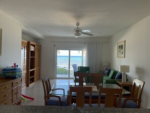 A living room with a view of the ocean, a ceiling fan, a green sofa, and a dining area with a glass table.