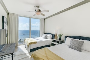A hotel room with two beds, a desk, a ceiling fan, and a large window offering a sea view.
