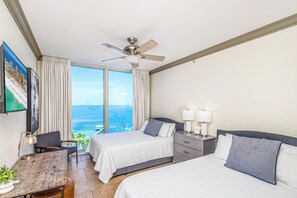 Ein Hotelzimmer mit Bett, Nachttischen, Schreibtisch, Stuhl, Deckenventilator und einem großen Fenster mit Meerblick.