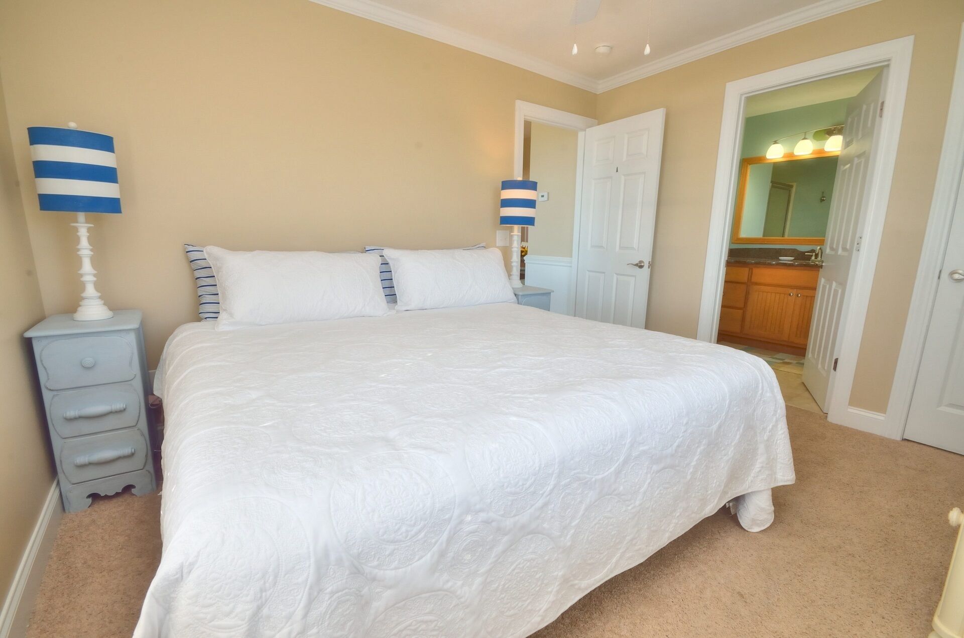 A neatly made bed with white bedding, a nightstand with a lamp, and a door leading to a bathroom with a mirror and lights.