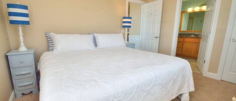 A neatly made bed with white bedding, a nightstand with a lamp, and a door leading to a bathroom with a mirror and lights.