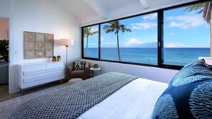 A bedroom with a large window, a bed with patterned bedding, a nightstand, a lamp, and a decorative wall piece. - GORGEOUS OCEANFRONT PENTHOUSE  (Lahaina)