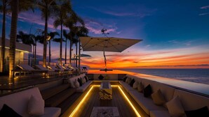 A sunset view from a deck with a sofa and lounge chairs, palm trees, and a large umbrella. - Pier 57 Luxury Escape 2BR, PV’s Best Rooftop! (Puerto Vallarta)