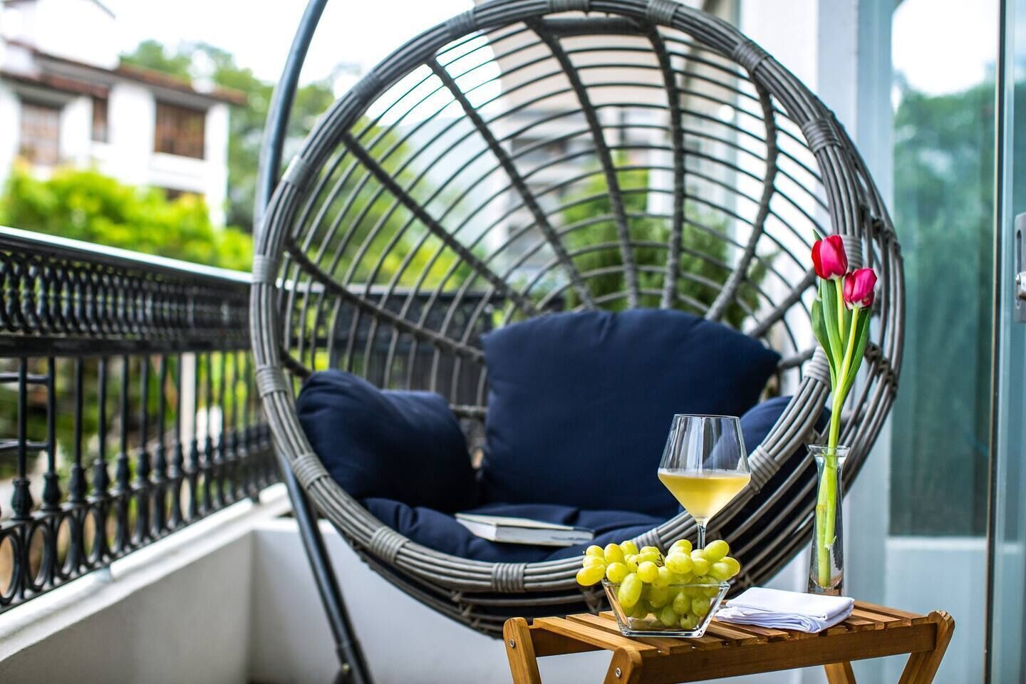 Une chaise en osier avec un coussin, un verre de vin blanc, un bol de raisins verts et un vase de tulipes rouges sur une petite table en bois.