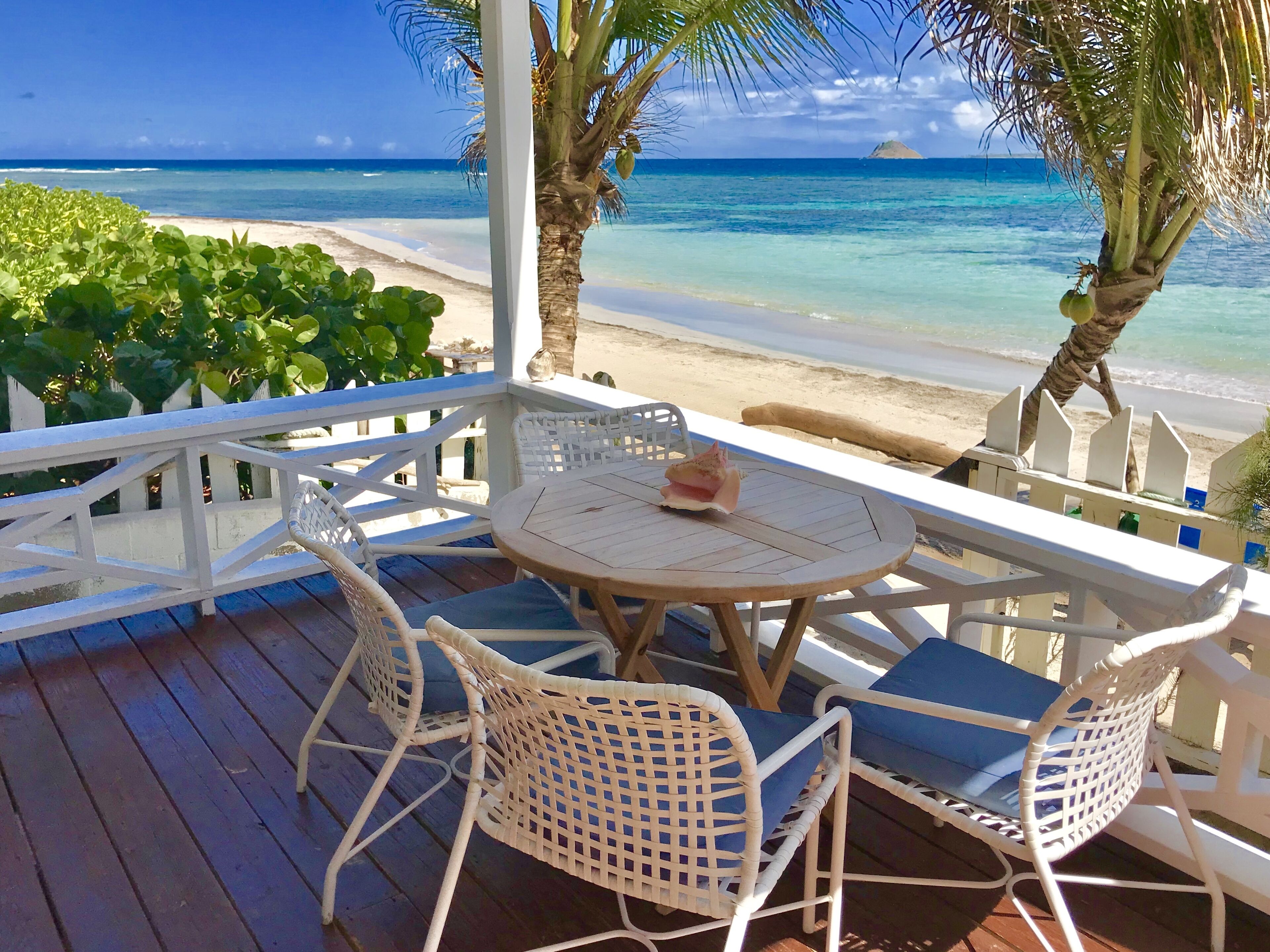 Terraza de madera con sillas de mimbre blanco y una mesa, con vista a una playa de aguas turquesas cristalinas y una isla distante.