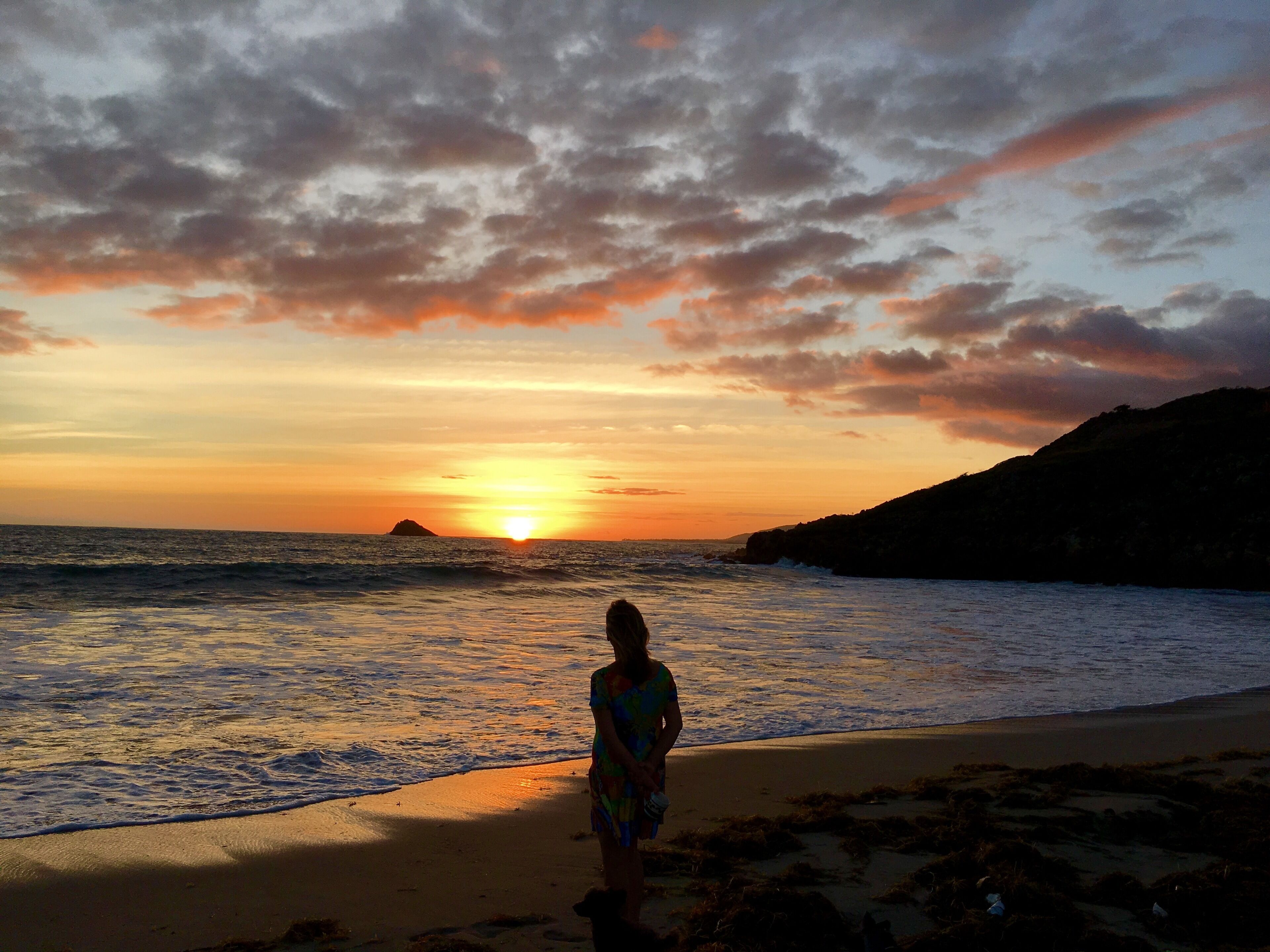A sunset over a beach with waves, a person standing facing the sunset, and a rocky coastline.