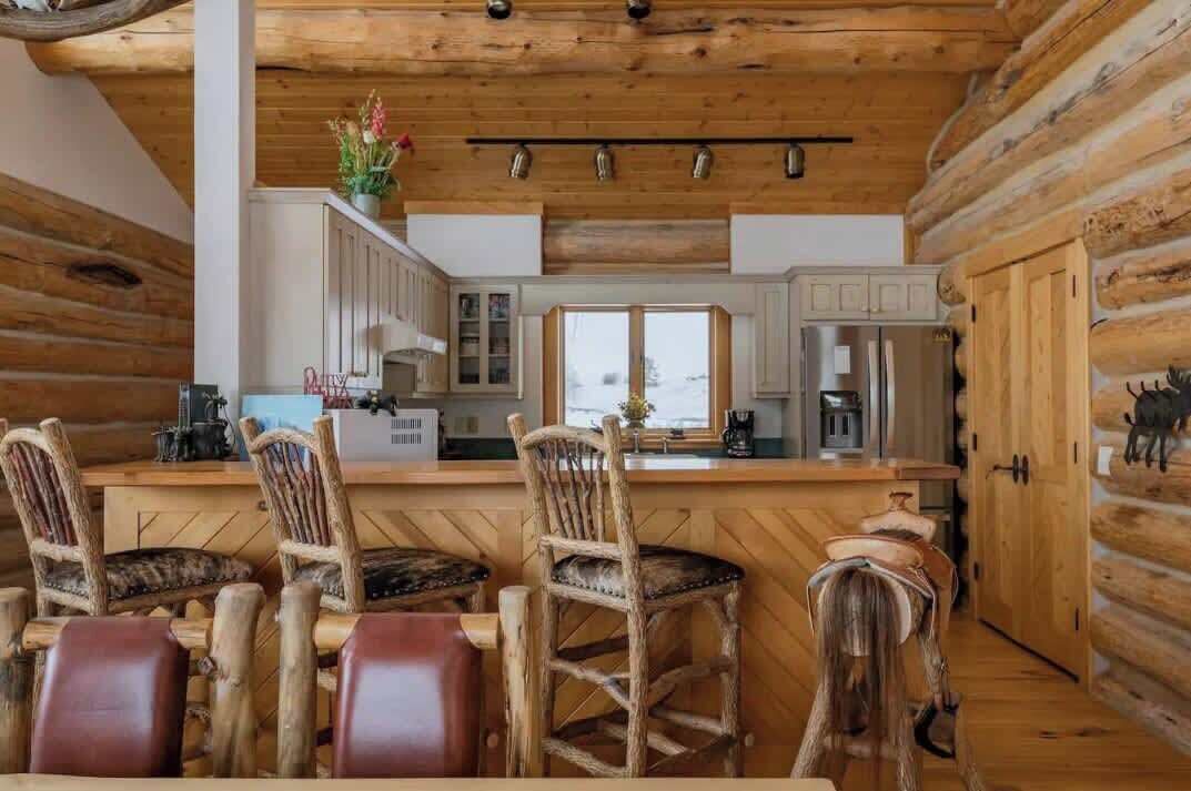 A rustic kitchen with wooden walls, a central island with bar stools, and a view of the snowy landscape.