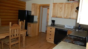 A compact kitchen with wooden cabinets, a stove, a dishwasher, and a dining area with a wooden table and chairs. - Cute 2/1 Cabin With Full Kitchen And Teton Views (Moran)