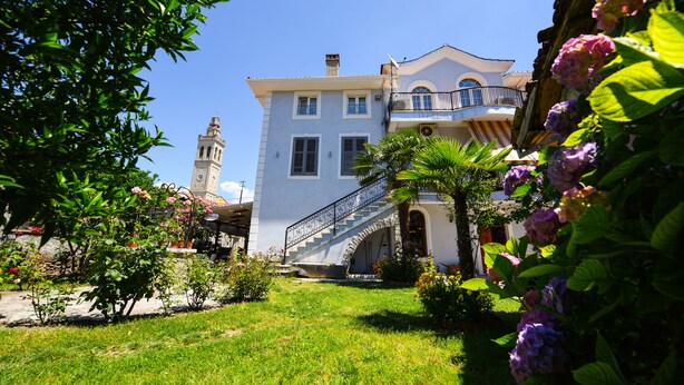 A two-story building with a balcony, a staircase, and a clock tower in the background. - Rose Garden Hotel (Shkodër)