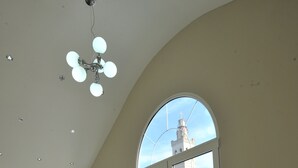 A spacious lobby with a modern chandelier, a staircase, and large arched windows offering views of a building with a clock tower. - Rose Garden Hotel (Shkodër)