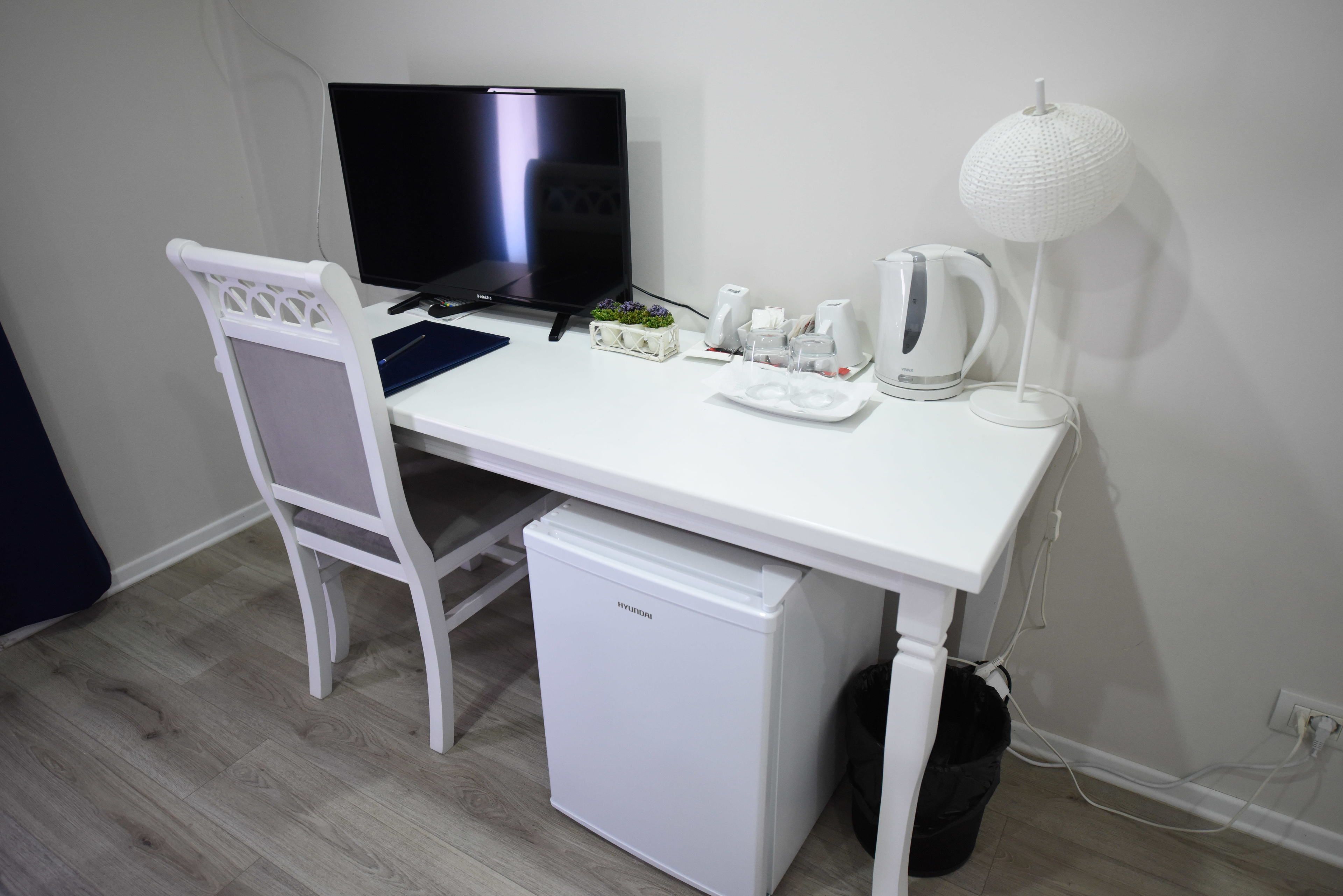 Un escritorio pequeño y blanco con un televisor, una lámpara blanca, un hervidor y un refrigerador.