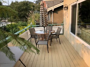 En træterrasse med et glasbord og stole, omgivet af grønne planter og udsigt over kvarteret.