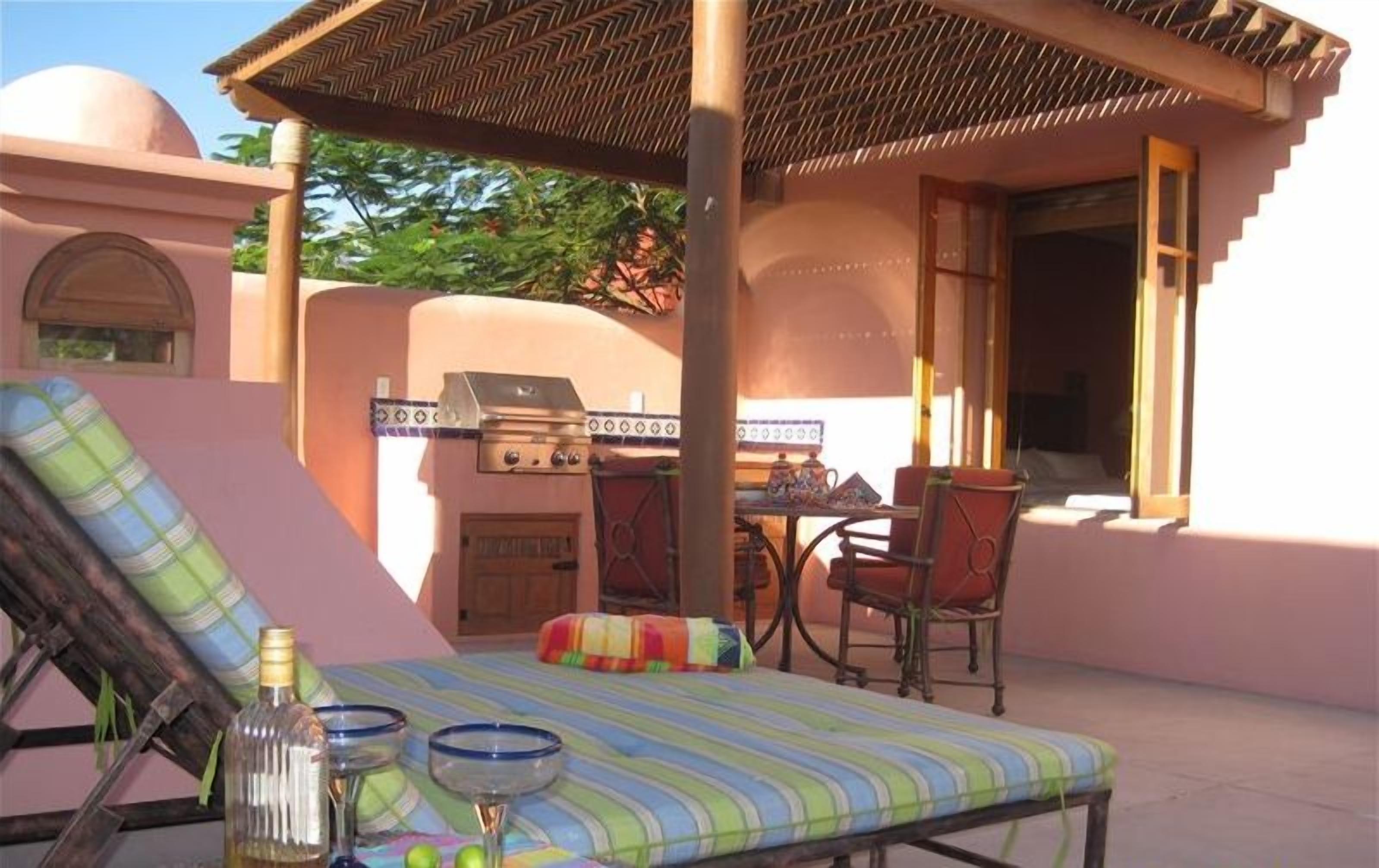 A sunlit outdoor seating area with a striped lounge chair, a table with a teapot, and a grill.