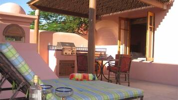 A sunlit outdoor seating area with a striped lounge chair, a table with a teapot, and a grill.