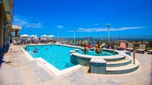 Zona de piscina con sillones de descanso y sombrillas, rodeada por un edificio de gran altura.