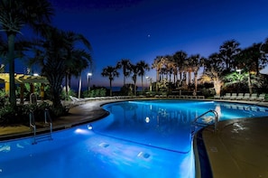 Une piscine éclairée la nuit, entourée de palmiers et de chaises longues.