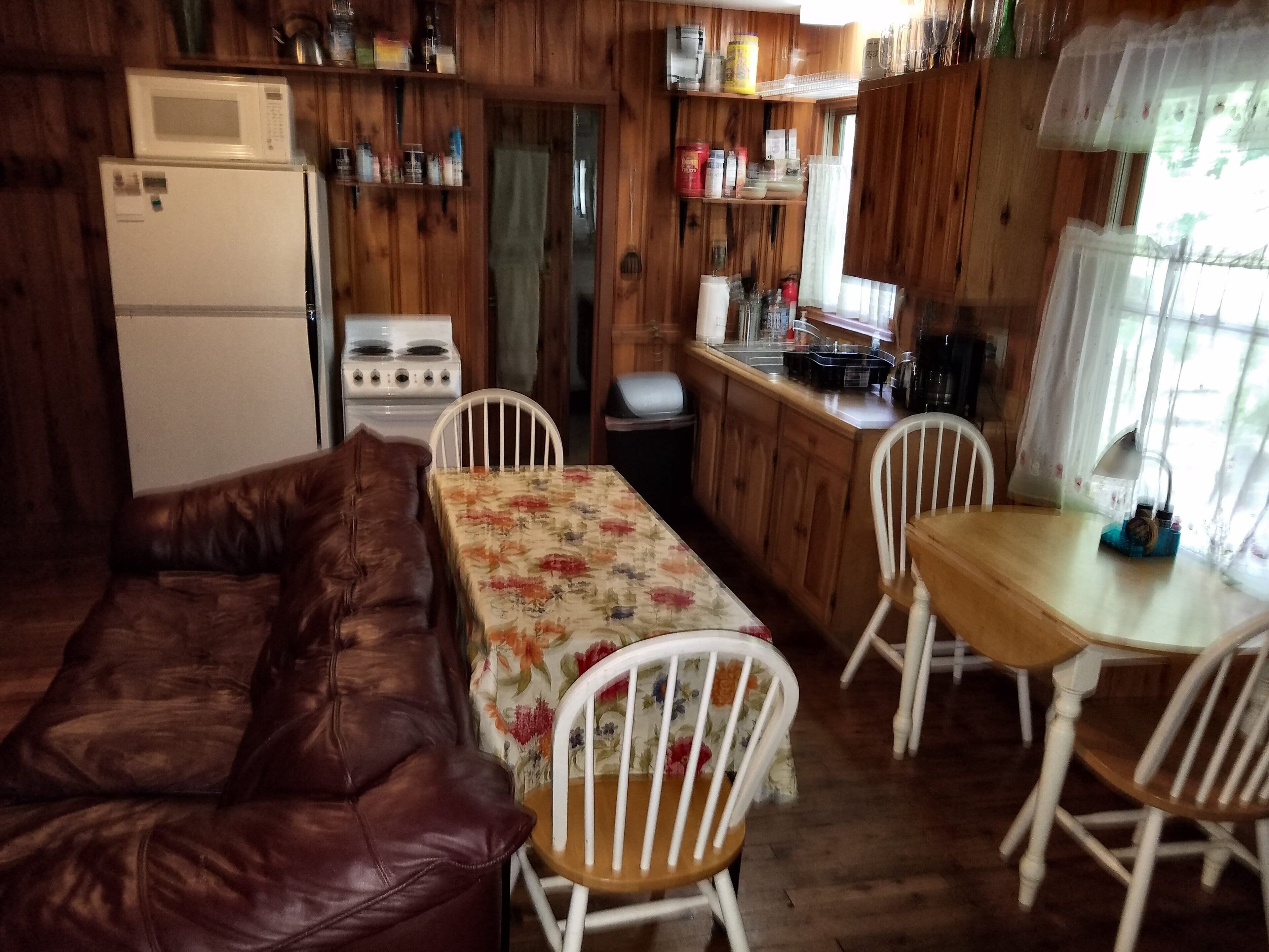Une cuisine chaleureuse, aménagée dans le style d’un chalet, avec des murs en bois, une table à manger entourée de chaises, un canapé en cuir marron, un réfrigérateur, une cuisinière et divers ustensiles de cuisine rangés sur des étagères.