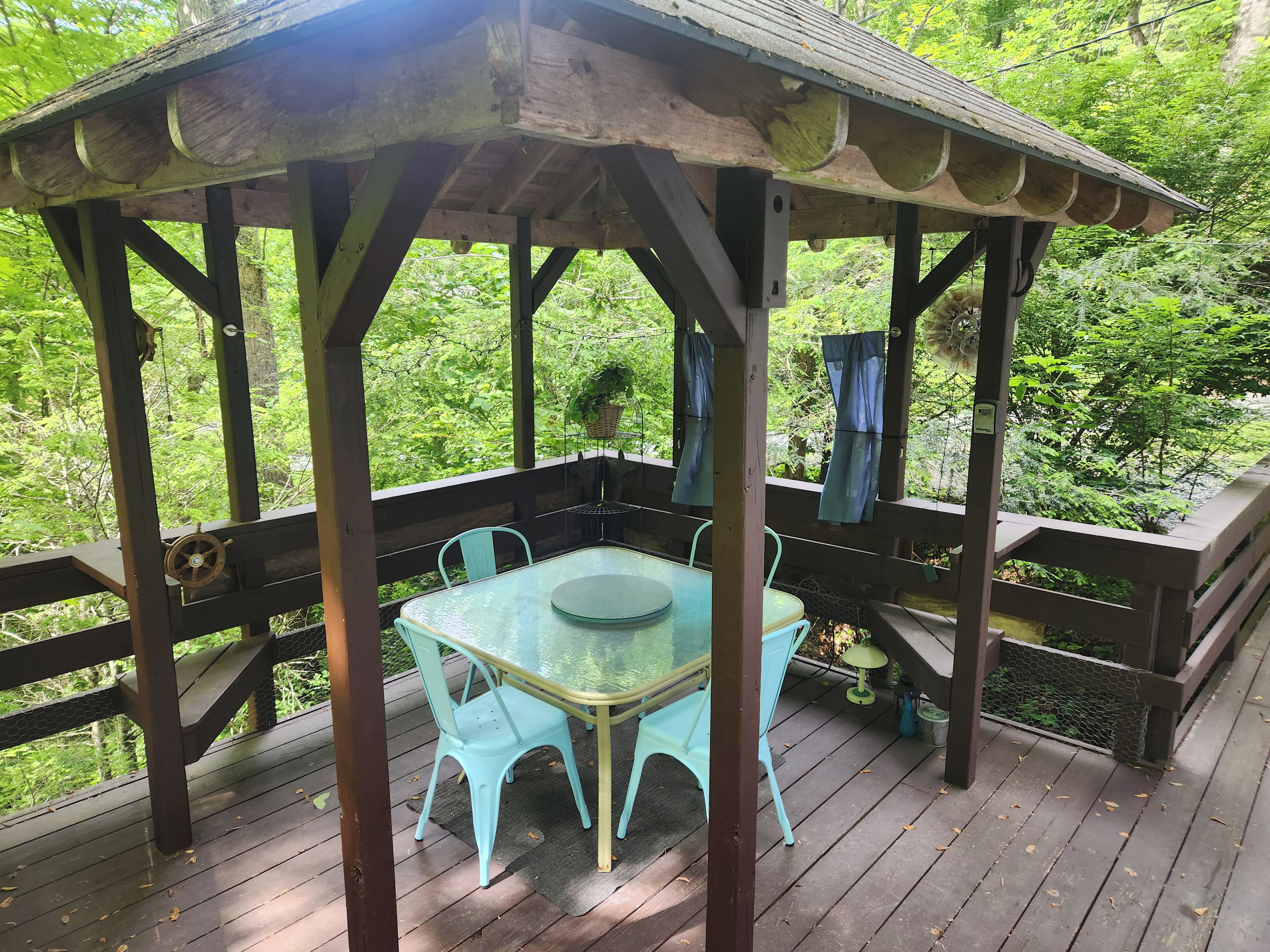 Eine hölzerne Laube mit einem Glastisch und Stühlen, umgeben von einer Holzterrasse und üppigem Grün.