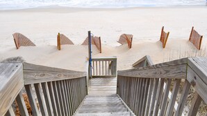 Una escalera de madera que baja hacia una playa arenosa, con una cerca y un portón de madera.