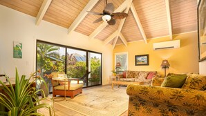 Un salon spacieux avec un plafond haut, des poutres en bois, une grande fenêtre donnant sur un paysage verdoyant, un canapé à motifs tropicaux et un ventilateur de plafond.