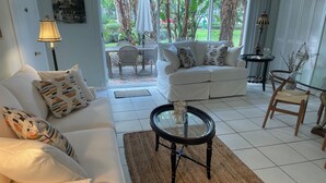 Ein modernes Wohnzimmer mit einem weißen Sofa, einem Glastisch und Blick auf einen Garten durch große Fenster.