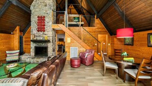 A spacious loft with a stone fireplace, wooden beams, a dining area, and a staircase leading to an upper level. - Honey Bear– Unique A-Frame Chalet w/ Mt LeConte View & Hot Tub in Gatlinburg (Gatlinburg)
