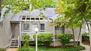 Un immeuble résidentiel à la façade grise, avec le numéro « 95 » bien visible sur la porte, un petit porche et un jardin bien entretenu, agrémenté de diverses plantes et d’un chemin.