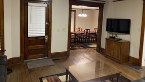 Un salon doté d’un plancher en bois, un coin repas avec une table et des chaises rouges, un téléviseur posé sur un meuble en bois, et une porte avec une fenêtre.