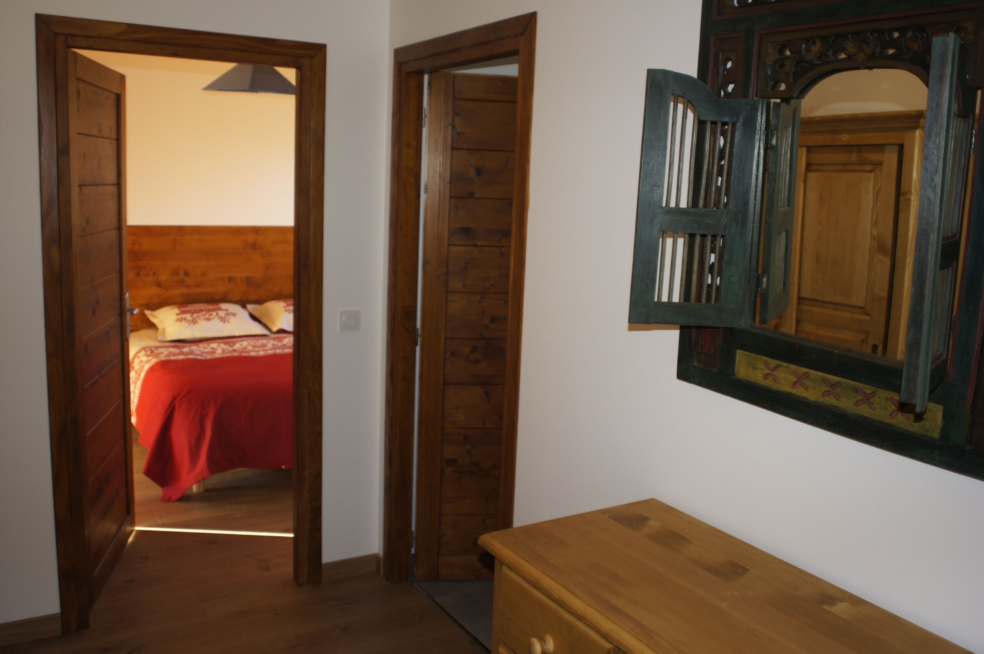Una stanza con una porta in legno che conduce a un letto con copriletto rosso e cuscini bianchi. Si nota un comò in legno e uno specchio con cornice decorativa appeso al muro.