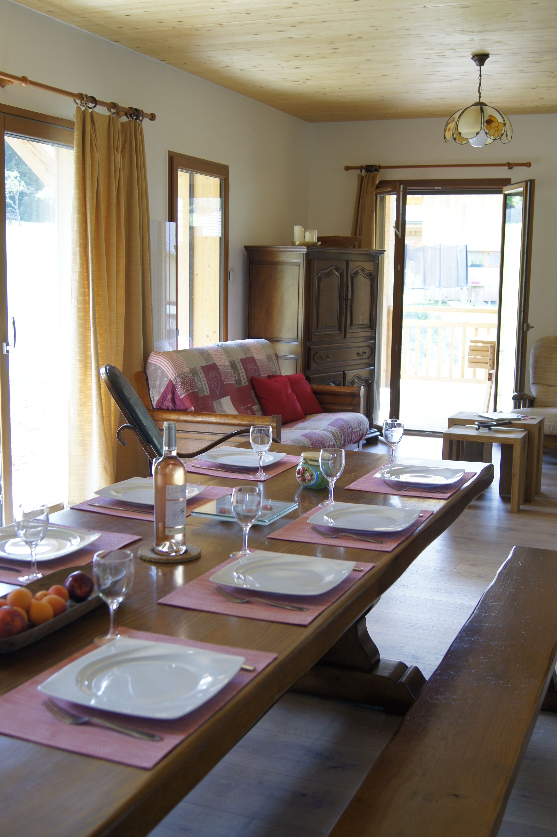 A dining area with a long wooden table set for four, a wooden bench, and a view of a balcony through an open door.