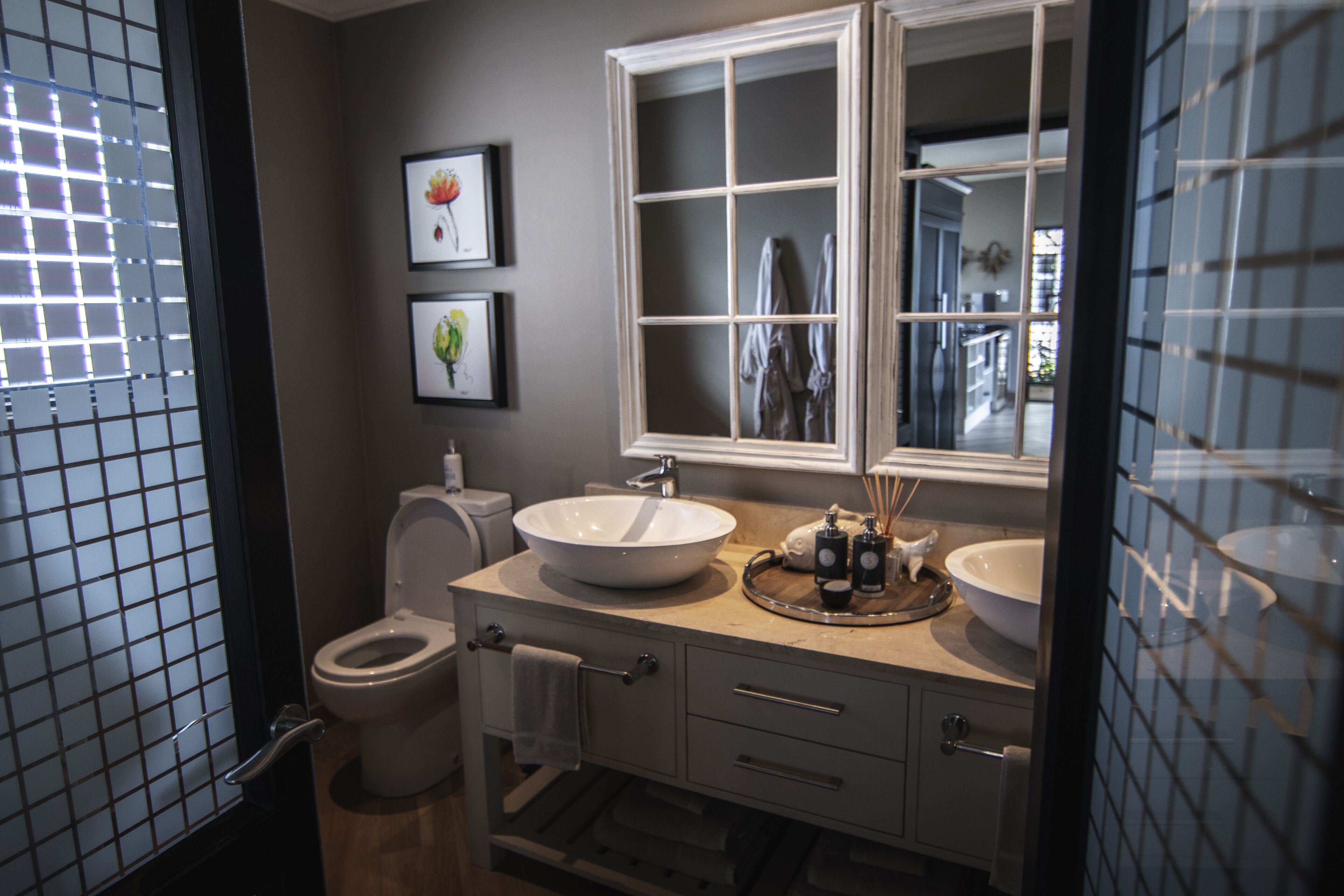 Ein Badezimmer mit Toilette, Waschbecken und Spiegel. An der Wand hängen Bilder in Rahmen und es gibt ein Fenster mit Jalousien.