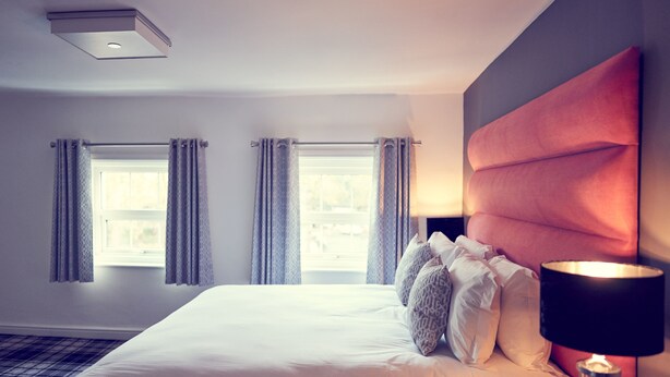 Une chambre d’hôtel avec un grand lit, deux fenêtres avec des rideaux, une tête de lit moderne et une lampe de chevet.