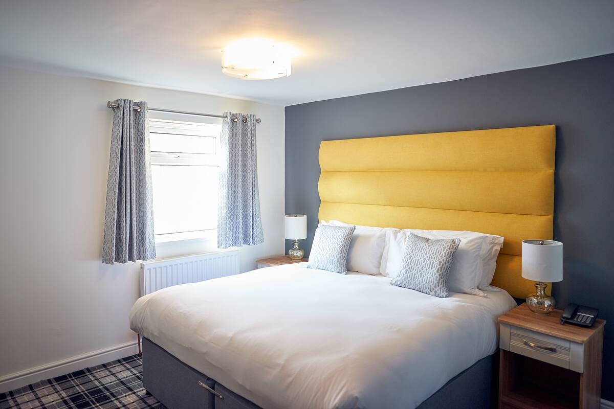 A neatly made bed with a yellow headboard, a bedside table with a lamp, and a window with curtains.