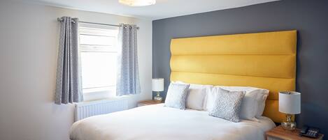 A neatly made bed with a yellow headboard, a bedside table with a lamp, and a window with curtains.