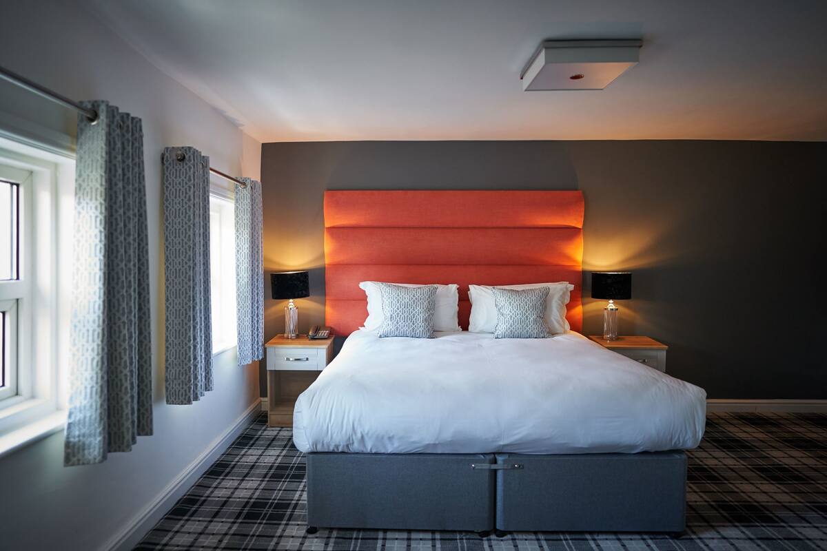 A modern hotel room with a large bed, two bedside tables with lamps, a headboard with built-in lighting, and a window with patterned curtains.