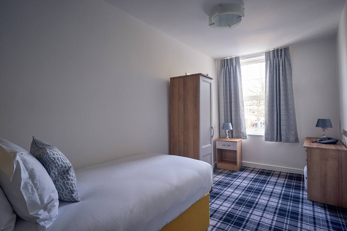 A bedroom with a bed, a wardrobe, a desk, a lamp, and a window with curtains.